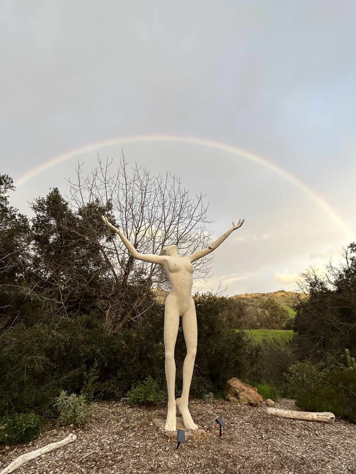 A sculpture of a figure with arms outstretched toward a rainbow arcing over a canyon landscape